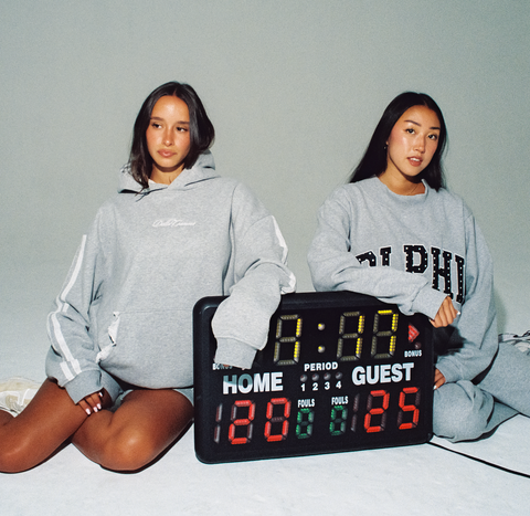 Two people wearing gray sorority hoodies with a digital scoreboard between them on a light gray background.