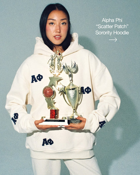 Sorority girl wearing a white hoodie with Alpha Phi sorority symbols, holding trophies against a light blue background. Link leads to Alpha Phi "Scatter Patch" Sorority Hoodie